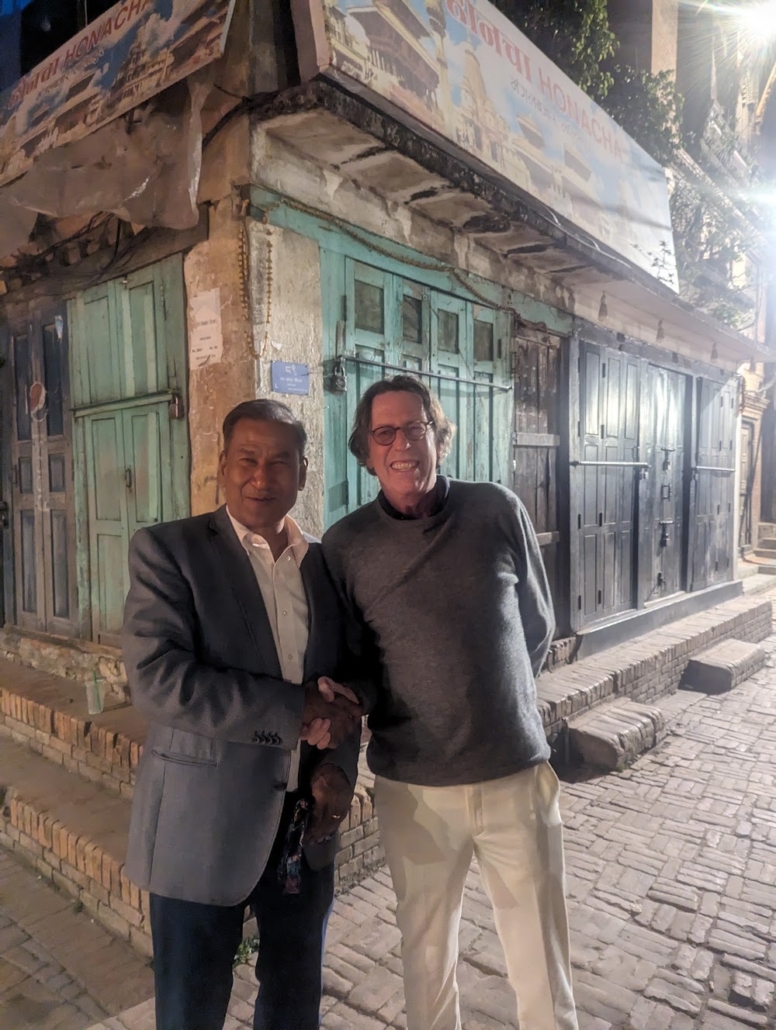 Rtn. Ewerhard Engels and Rtn. Bijaya Mallapaty shake hands on a brick-paved street in Patan’s heritage area at night, 15 April 2024.